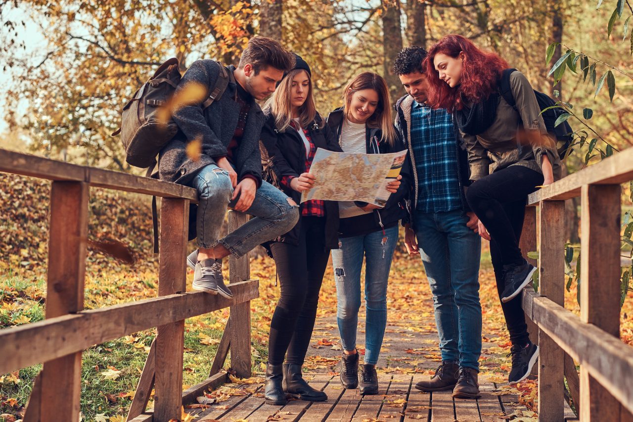 group of young friends hiking in autumn colorful forest looking at map and planning hike e1699012191269.jpg
